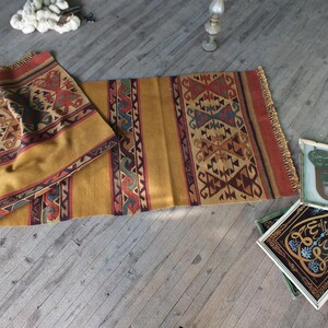 May include: A traditional Turkish kilim rug with a geometric pattern in shades of red, brown, blue, and yellow. The rug is folded in half and lying on a wooden floor.