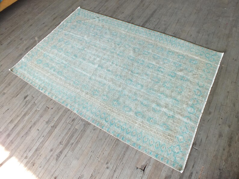 K&ouml;nnte beinhalten: Ein hellblauer und wei&szlig; gemusterter Teppich mit einem verblassten, Vintage-Look. Der Teppich hat einen Rand aus hellblauen und wei&szlig;en geometrischen Mustern.