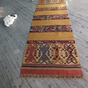 May include: A long, rectangular rug with a geometric pattern in shades of red, blue, green, and brown. The rug has a fringe border and is made of wool.