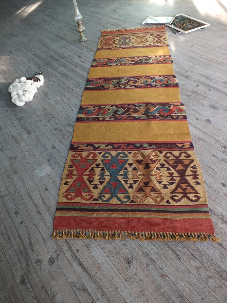May include: A long, rectangular rug with a geometric pattern in shades of red, blue, green, and brown. The rug has a fringe border and is made of wool.
