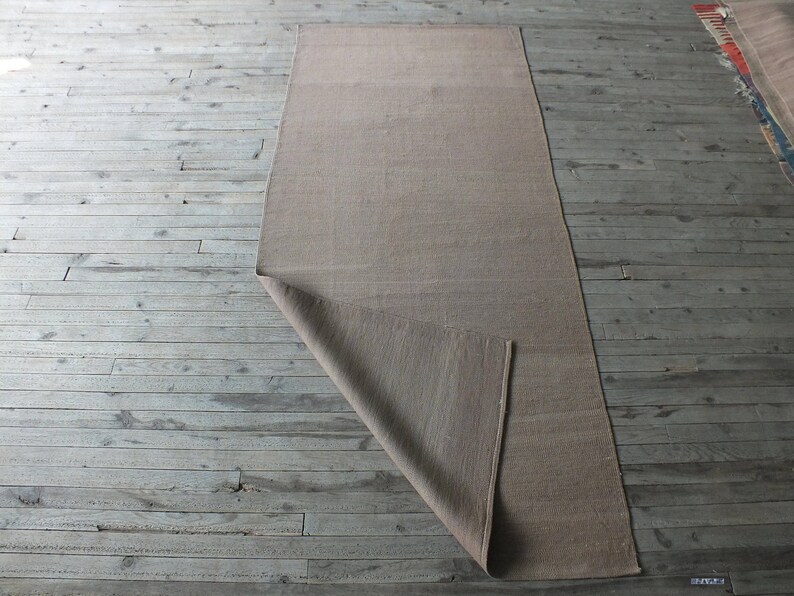 May include: A beige rug is folded in half and laid out on a wooden floor. The rug is made of a natural material and has a subtle texture.