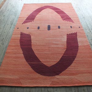 May include: A large, rectangular rug with a unique design. The rug is a light orange color with a large, dark red, U-shaped design in the center. The design is reminiscent of an archway or a tunnel.