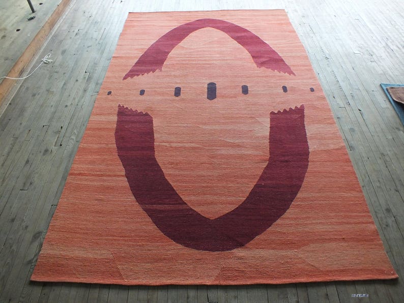 May include: A large, rectangular rug with a unique design. The rug is a light orange color with a large, dark red, U-shaped design in the center. The design is reminiscent of an archway or a tunnel.