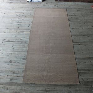 May include: A light brown rectangular rug with a subtle woven texture. The rug is laid out on a wooden floor.