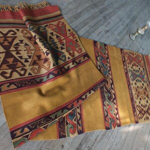 May include: A traditional Turkish kilim rug with a geometric pattern in shades of brown, red, blue, and green. The rug has a fringe on both ends and is made of wool.