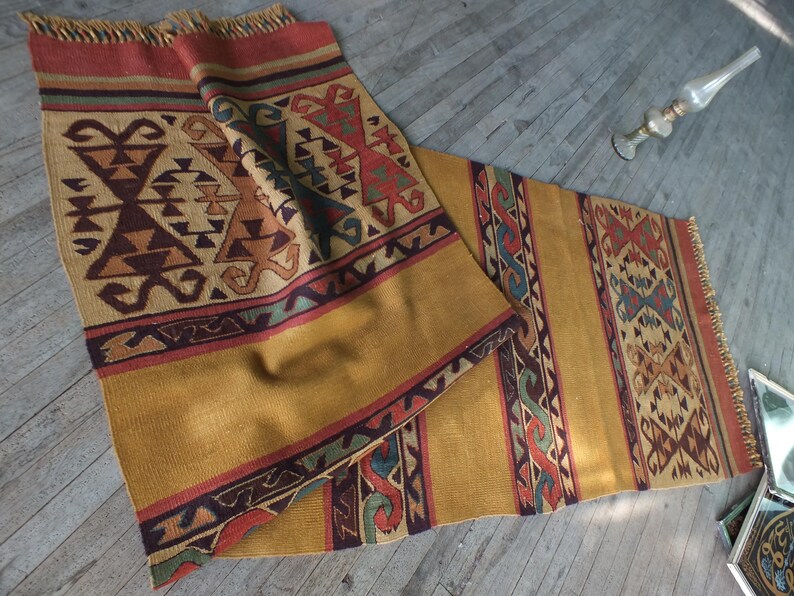 May include: A traditional Turkish kilim rug with a geometric pattern in shades of brown, red, blue, and green. The rug has a fringe on both ends and is made of wool.