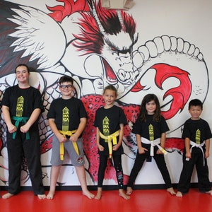 以下が含まれることがあります： 黄色と緑のベルトをつけた黒い武道着を着た6人のグループが、赤と黒の大きな龍の壁画のある白い壁の前に立っています。壁画には、赤い尾と白い体をした龍が描かれています。みんな笑顔でカメラを見ています。