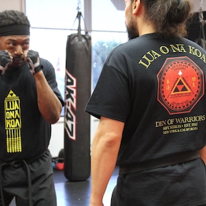May include: Two people wearing black martial arts uniforms with yellow and gold logos. The person on the left is wearing black gloves and has their hands up in a defensive position. The person on the right is facing away from the camera. The text on the back of the shirt says "LUA O NA KOA" and "DEV OF WARRIORS".