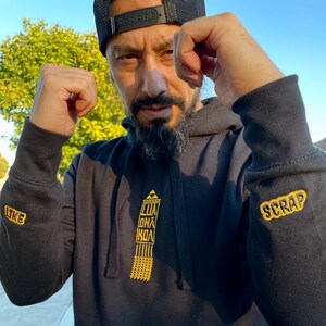Könnte beinhalten: Schwarzer Kapuzenpullover mit Fronttasche und goldfarbenem, gesticktem Text und Design. Der linke Ärmel trägt die Aufschrift "LIKE" und der rechte Ärmel "SCRAP". Auf der Vorderseite befindet sich ein goldenes Design und der Text "LUA KOA".