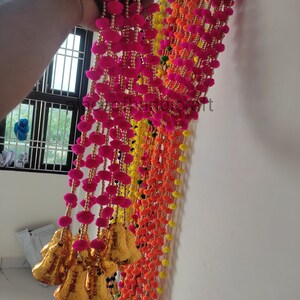 May include: A colourful garland of pink, orange and yellow pom-poms with gold bells hanging from the bottom. The garland is made of yarn and beads.