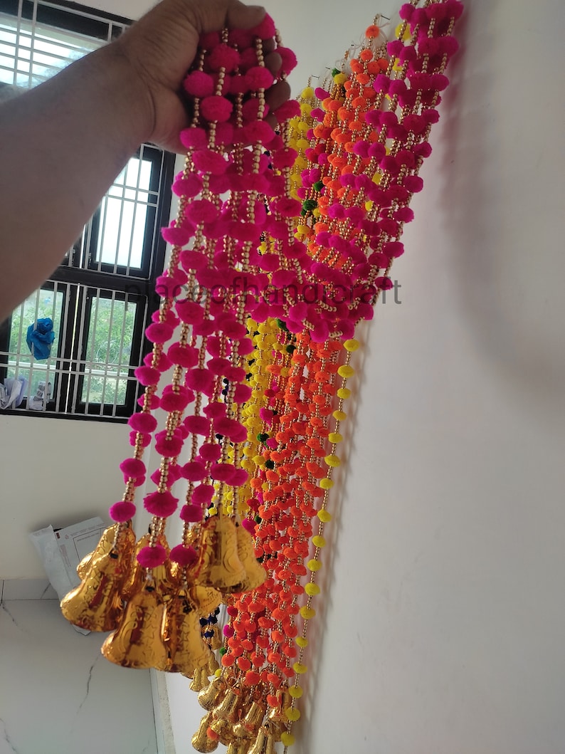 May include: A colourful garland of pink, orange and yellow pom-poms with gold bells hanging from the bottom. The garland is made of yarn and beads.