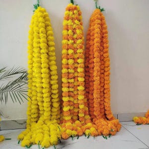 May include: Three long vertical garlands of artificial marigold flowers hanging against a wall. The left garland is bright yellow, the middle garland features a mix of yellow and orange blossoms, and the right garland is a solid deep orange color.