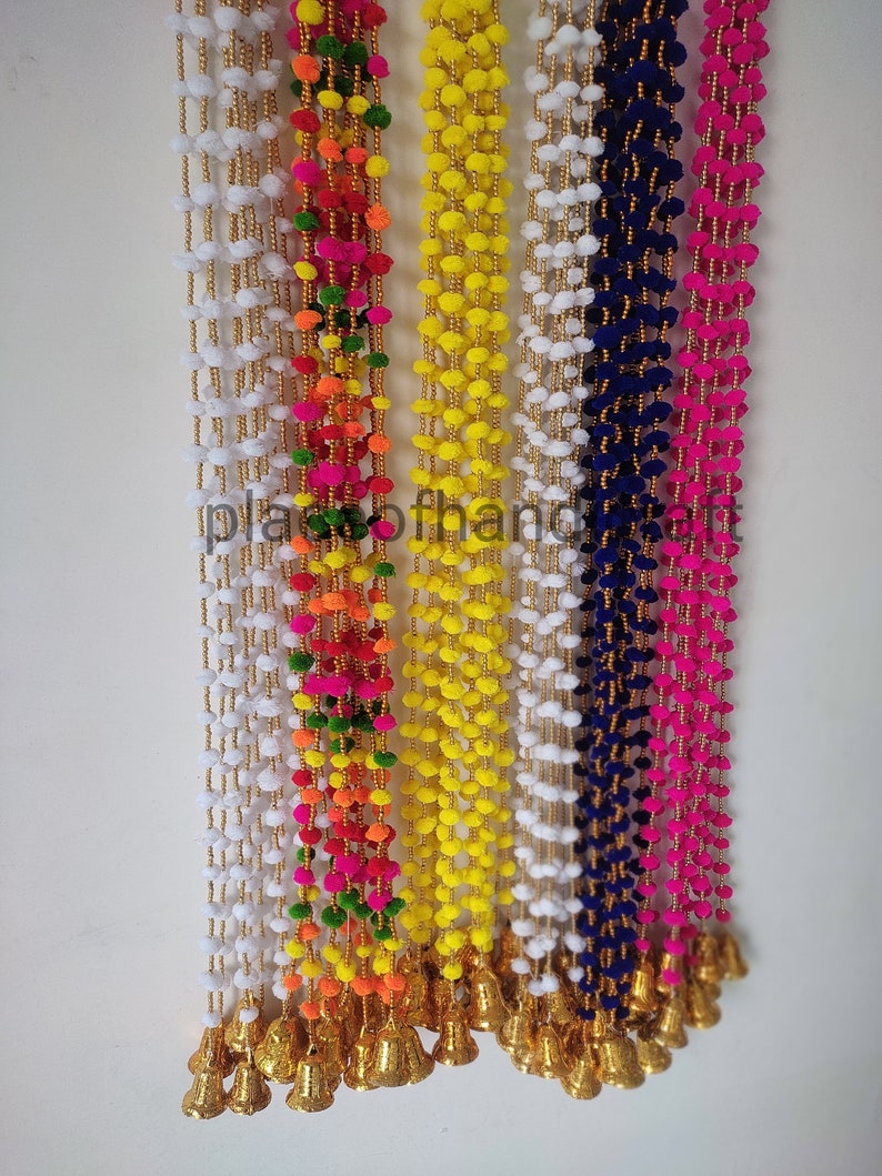 May include: A set of six colourful pom-pom garlands with gold bells at the bottom. The garlands are made of white, yellow, pink, blue, green, and orange pom-poms.