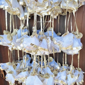 May include: A group of white tulle bags with gold trim and gold tassels hanging from a wooden surface. The bags are tied with gold ribbon and have a gold trim around the top. The bags are all the same size and shape. "place of handicraft" is written on the wooden surface.