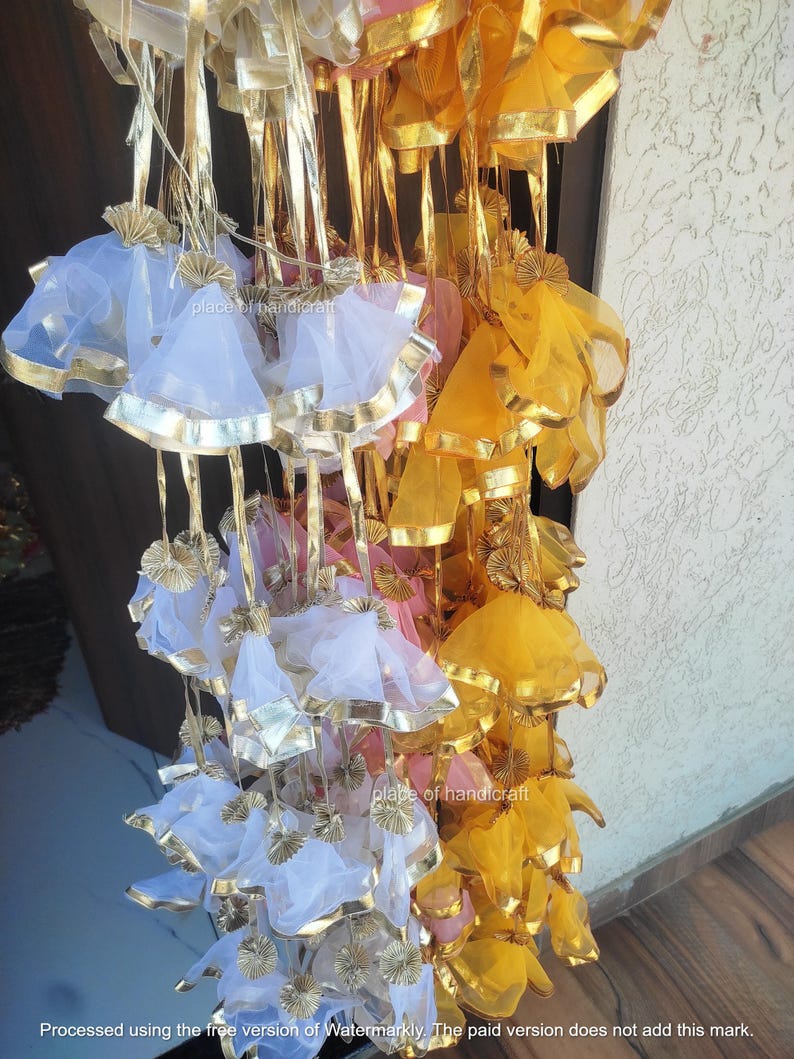 May include: A collection of decorative hanging pouches made of sheer fabric in white, yellow, and pink. The pouches have gold trim and are strung together with gold ribbon.
