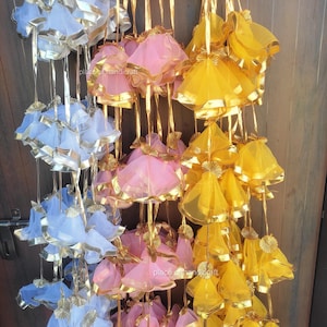 May include: Rows of decorative hanging ornaments in shades of blue, pink, and yellow. Each ornament features a bell shape with gold trim and a gold string for hanging. The ornaments are made of a sheer fabric.