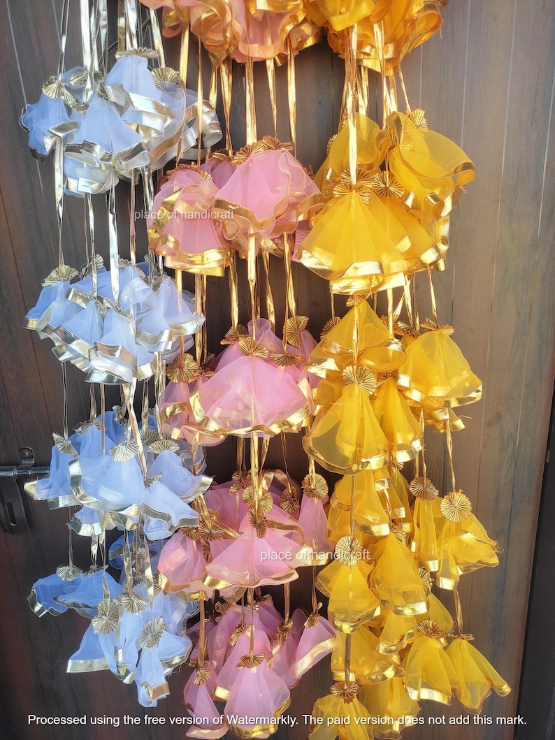 May include: A string of decorative pouches made of sheer fabric in blue, pink, and yellow. Each pouch has a gold ribbon trim and is tied with a gold ribbon.