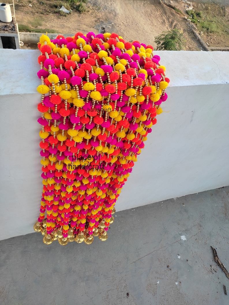 May include: A long garland of colourful pom-poms in pink, yellow, and orange. The pom-poms are strung together with gold beads and have gold bells at the bottom.