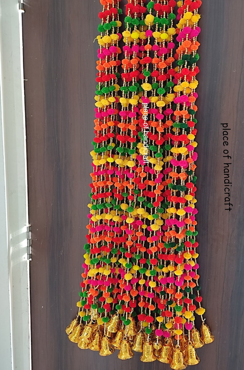 May include: Multicoloured pom-pom garland with gold bells. The garland is made of small, fluffy balls in shades of pink, orange, yellow, and green. The garland is strung together with gold beads and has a row of gold bells at the bottom.