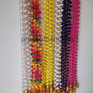 May include: A set of six colorful pom-pom garlands with gold bells at the bottom. The garlands are made of white, yellow, pink, blue, green, and orange pom-poms.