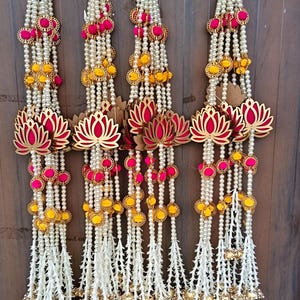 May include: Four decorative wall hangings featuring strings of white faux pearls interspersed with bright pink and yellow fabric pom-poms. Each strand includes a central cutout lotus flower ornament and ends with small gold-toned metal bells.