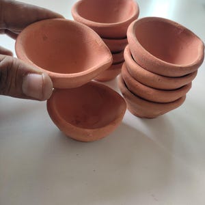 Könnte beinhalten: Mehrere kleine Öllampen aus Terrakotta-Ton sind gestapelt und werden in einer Hand gehalten. Die Lampen haben eine flache, abgerundete Schalenform mit einem kleinen Ausguss. Die Lampen haben einen warmen, erdigen Ton.