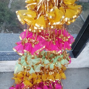 May include: A large stack of colorful fabric gift bags with gold trim. The bags are made of sheer fabric and have a drawstring closure. The bags are arranged in a pyramid shape, with the yellow bags at the top, the pink bags in the middle, and the green bags at the bottom.