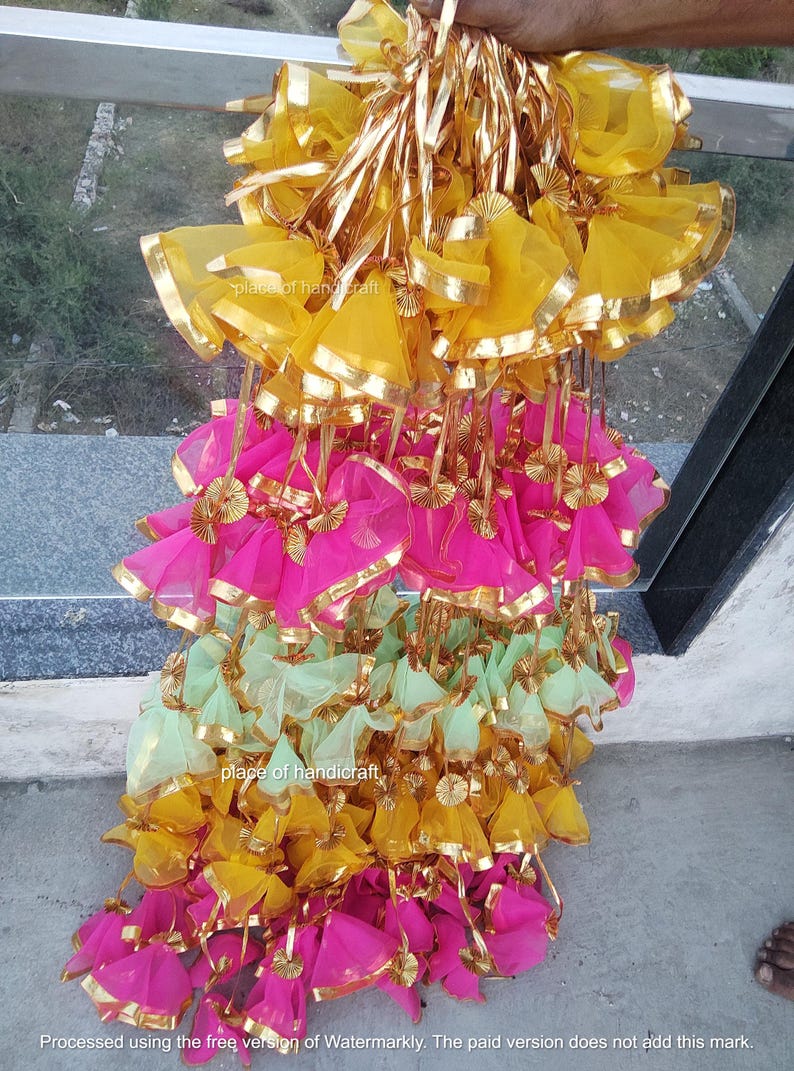 May include: A large stack of colorful fabric gift bags with gold trim. The bags are made of sheer fabric and have a drawstring closure. The bags are arranged in a pyramid shape, with the yellow bags at the top, the pink bags in the middle, and the green bags at the bottom.