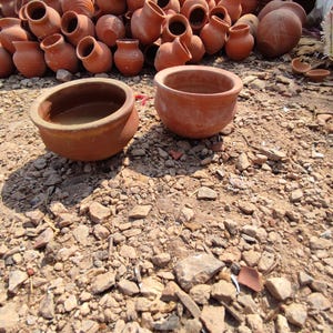 May include: A collection of terracotta pots and vessels, varying in size and shape, are displayed outdoors. The unglazed pottery has a reddish-brown hue, with some pots stacked in the background and others scattered in the foreground on a bed of gravel and dirt.
