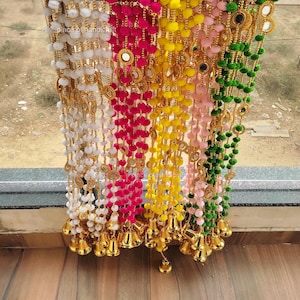 以下が含まれることがあります： 白、ピンク、黄色、緑のポンポンが付いた装飾的なドアハンギングが4本。各ストランドには、小さな丸い鏡と金のベルが付いています。