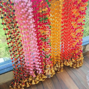 May include: Colorful pom-pom garlands in shades of pink, red, orange, and yellow hang vertically. Each strand is adorned with small, gold-colored bells at the bottom. The garlands are displayed against a natural background.