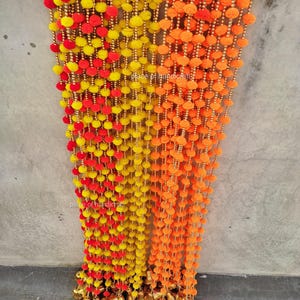 May include: Decorative garlands in yellow, red, and orange. The garlands are made of small, round, pom-pom-like elements strung together with gold-colored beads. The garlands hang vertically, with small gold bells at the bottom. The text "place of handicraft" is visible.