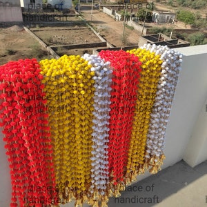 May include: A string of red, yellow and white pom-poms with gold bells at the bottom. The pom-poms are strung together with a gold thread. The string is hanging from a wall. The text "place of handicraft" is visible at the bottom of the image.
