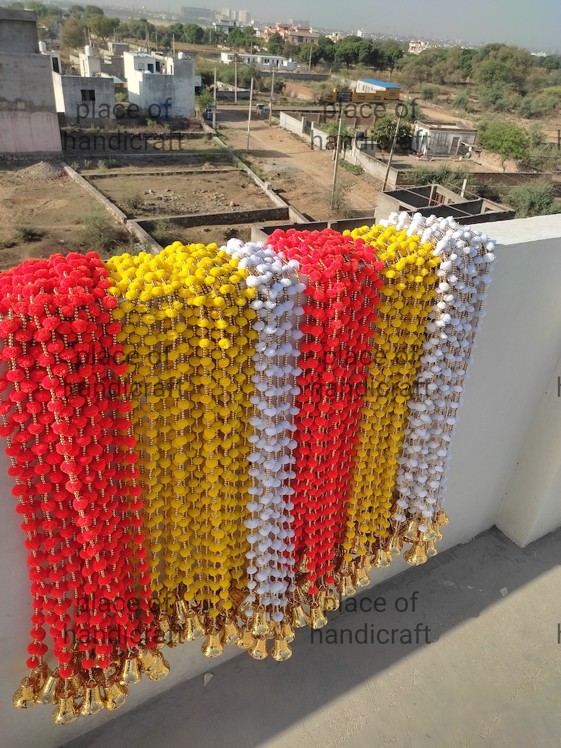 May include: A string of red, yellow and white pom-poms with gold bells at the bottom. The pom-poms are strung together with a gold thread. The string is hanging from a wall. The text "place of handicraft" is visible at the bottom of the image.