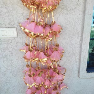May include: A string of pink and gold decorative tassels hanging from a white wall. The tassels are made of sheer fabric and have a gold trim. The tassels are arranged in a cascading pattern.