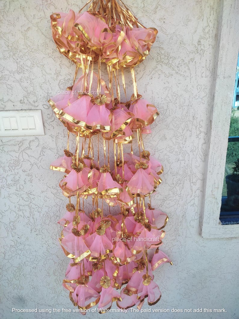 May include: A string of pink and gold decorative tassels hanging from a white wall. The tassels are made of sheer fabric and have a gold trim. The tassels are arranged in a cascading pattern.