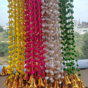 May include: A set of four decorative door hanging garlands with pom-poms in yellow, pink, white, and green colours. Each garland has gold bells at the bottom. The garlands are hanging from a window.