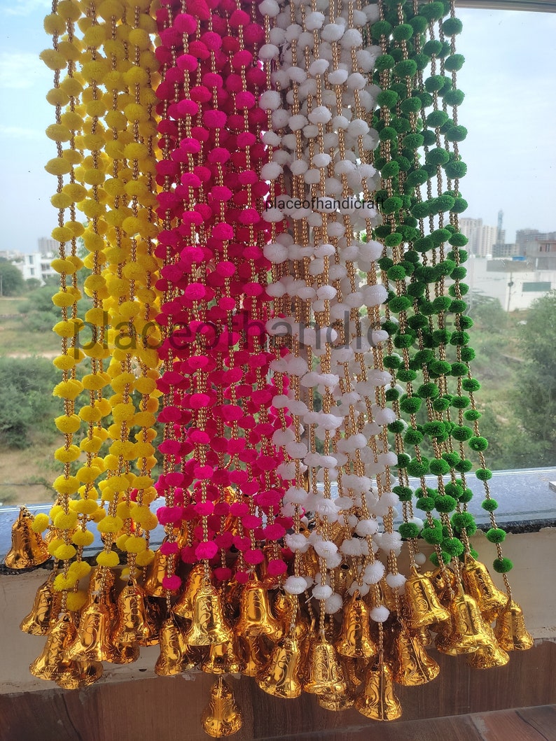 May include: A set of four decorative door hanging garlands with pom-poms in yellow, pink, white, and green colours. Each garland has gold bells at the bottom. The garlands are hanging from a window.