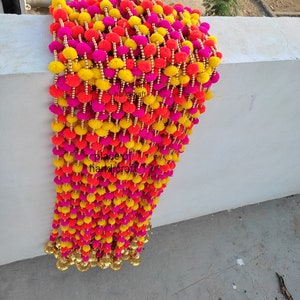 May include: A long garland of colorful pom-poms in pink, yellow, and orange. The pom-poms are strung together with gold beads and have gold bells at the bottom.