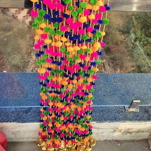 May include: A decorative hanging garland featuring multiple vertical strands of vibrant neon pink, bright orange, lime green, and deep blue pom-poms interspersed with small gold-colored beads, finished with metallic gold bells at the bottom ends.