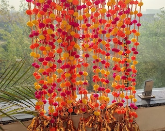 May include: A colorful garland made of pink, orange, and yellow pom-poms with gold bells hanging from the bottom. The garland is made of multiple strands and is hanging from a window.