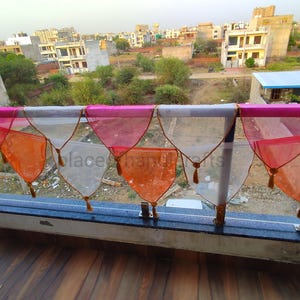 May include: A decorative banner with triangular, sheer fabric panels in shades of pink, orange, and white. Each panel is trimmed with a gold border and a tassel. The banner is strung along a metal railing, with a cityscape visible in the background.
