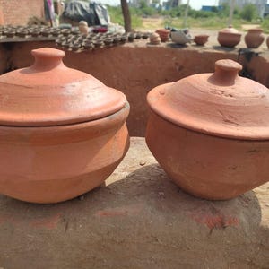 May include: Two terracotta clay pots with lids. The pots are a warm reddish-brown color and have a rounded shape. The lids have a small knob on top. The pots are sitting on a rough, earthy surface.
