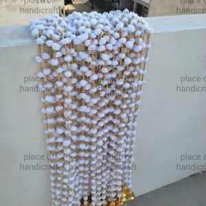 May include: A white pom-pom garland with gold beads and gold bells hanging from the bottom. The garland is hanging from a wall.