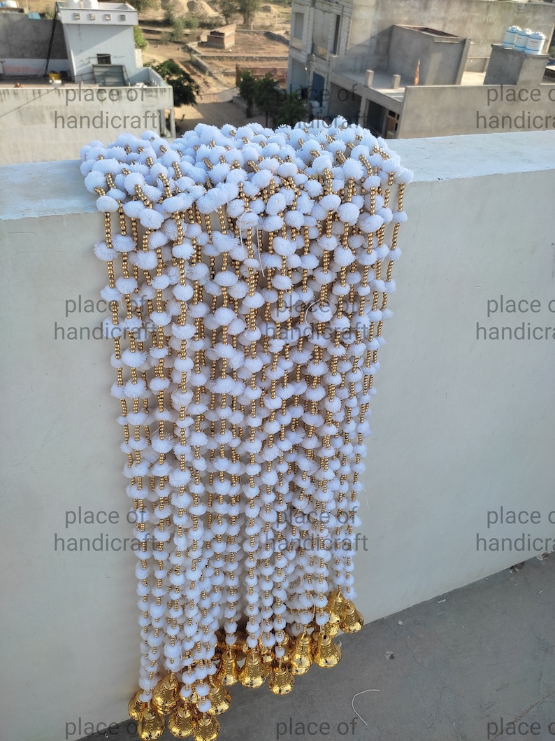 May include: A white pom-pom garland with gold beads and gold bells hanging from the bottom. The garland is hanging from a wall.