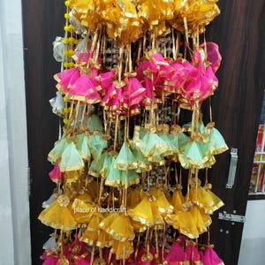 May include: A collection of colorful, decorative fabric ornaments in shades of yellow, pink, and green, hanging from a rod. Each ornament has a gold trim and is suspended by a gold string. The image is of a handicraft display.