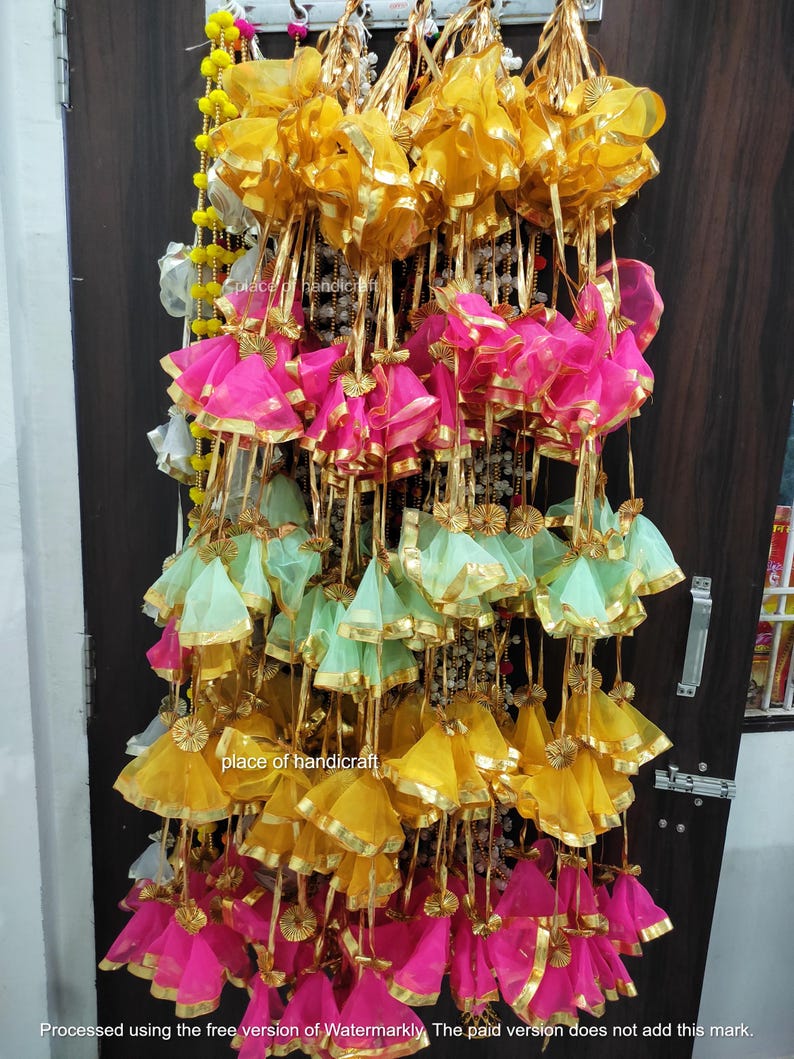 May include: A display of colorful fabric tassels with gold trim. The tassels are arranged in rows and are hanging from a wall. The tassels are made of a variety of colors, including pink, green, and yellow.