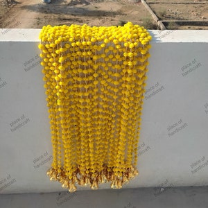 May include: A long strand of yellow pom-pom trim with gold beads and gold tassels. The trim is hanging from a white wall.