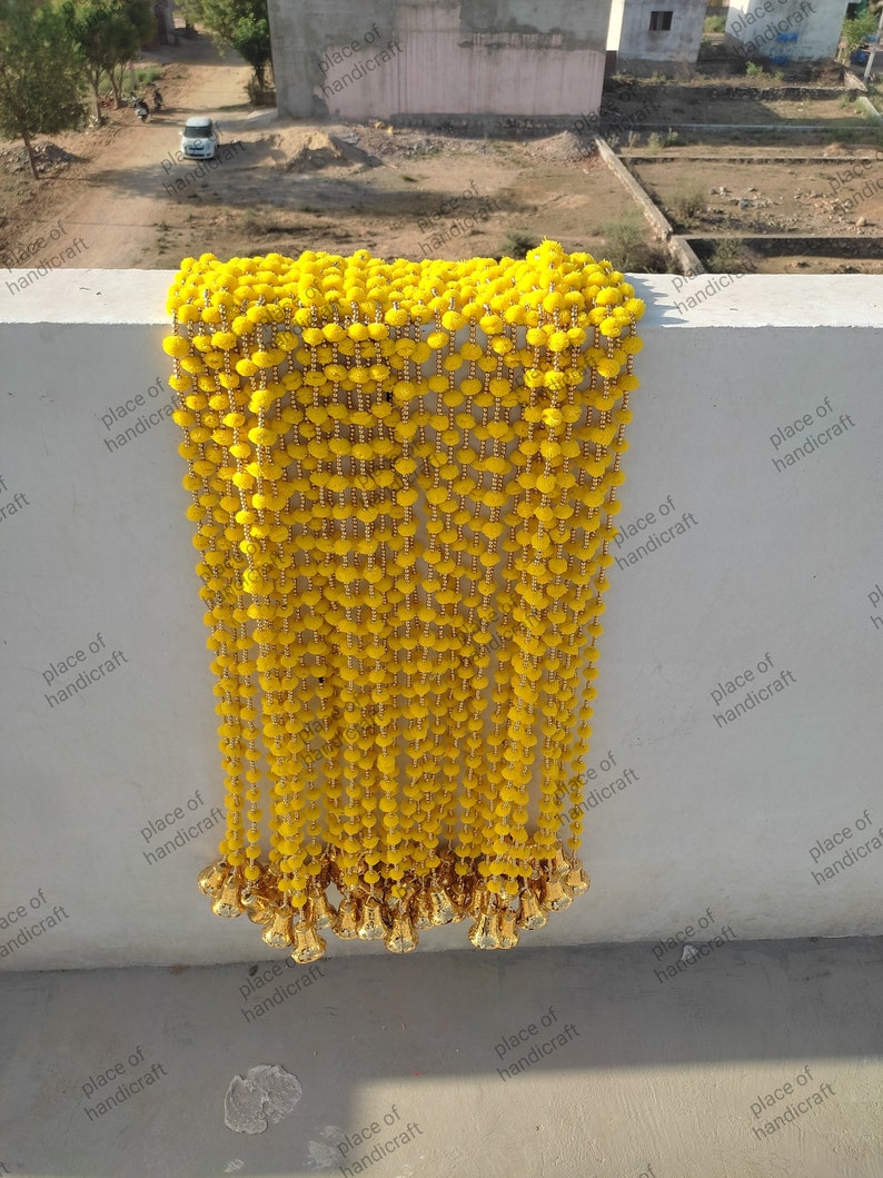 May include: A long strand of yellow pom-pom trim with gold beads and gold tassels. The trim is hanging from a white wall.