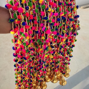 May include: A colorful garland of pom-poms with gold beads and bells. The garland is made of multi-colored yarn and is strung together with gold beads. The garland is decorated with gold bells at the bottom.